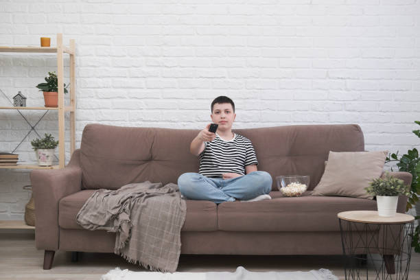 istock A teen boy is sitting on the sofa, eating pop corn, holding control panel and watching tv in light living room. 1477851390