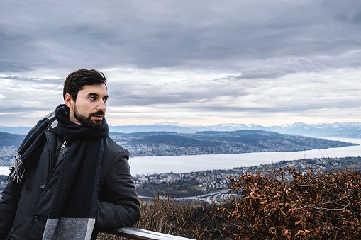 Young caucasian man with heavy clothing watching over Zurich lake from balcony on a height under overcast winter sky