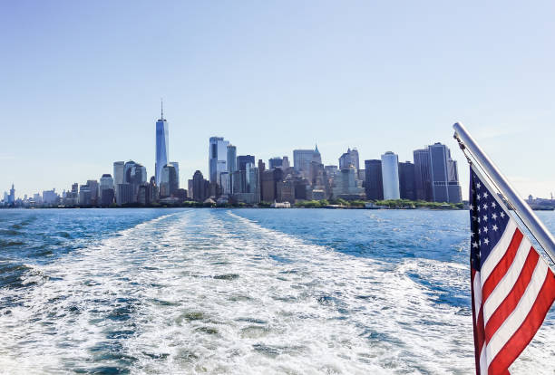 風光明媚なニューヨーク市のフェリー旅行。ニューヨークのスカイラインと風景。アメリカの国旗。建物、水、青い空。海の波。ボートまたはヨット。都市の川の風景。マンハッタンの観光� - lower downtown ストックフォトと画像