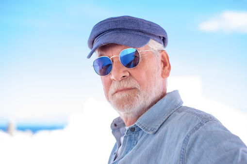 Portrait of senior man looking at camera. Face of a serious old man wearing blue sunglasses and cap outdoor. Retired relaxed man with white hair and beard in denim shirt
