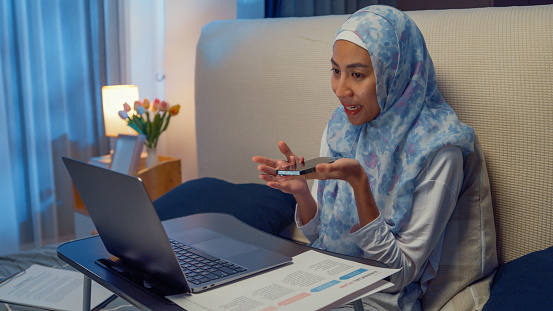 Happy young muslim woman sit on bed with computer laptop on folding laptop table and use smartphone giving voice commands to virtual assistant in bedroom at home at night. Work from home concept.