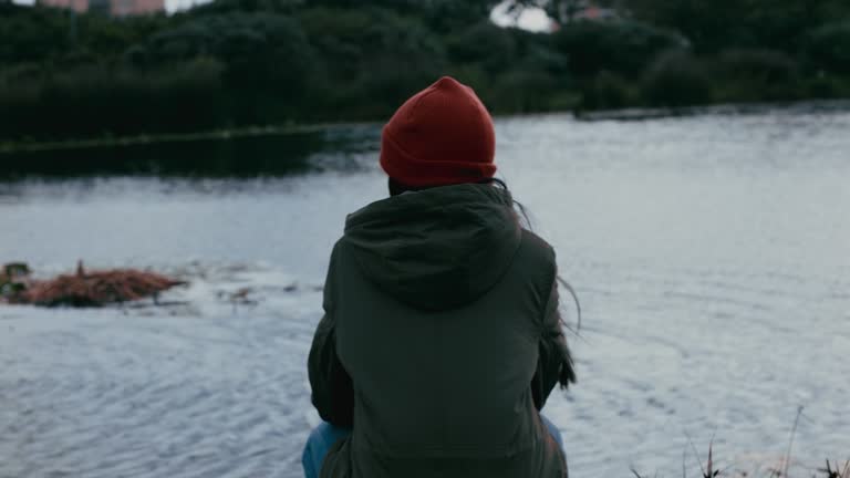 lonely young woman sitting in park enjoying peaceful natural pond teenage girl feeling depressed on cloudy day wind blowing hair