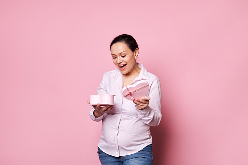 Smiling happy multi-ethnic pregnant woman, wearing casual white shirt and blue denim jeans, expressing happiness and positive emotion, rejoicing at present for Mother's Day on pink isolated background