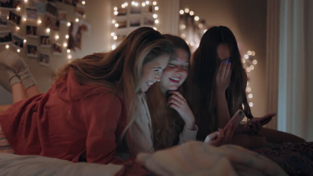 happy teenage girls lying on bed using smartphone watching online entertainment laughing having fun sleep over on weekend