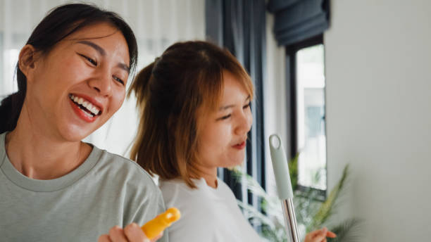 Closeup of young Asian lesbian couples having fun during cleaning house and singing with mop live online screen IG reel tiktok at living room. Dancing and singing in a cozy room concept. primer plano de jóvenes parejas de lesbianas asiáticas divirtiéndose durante la limpieza de la casa y cantando con la fregona en vivo en línea de la pantalla ig reel tiktok en la sala de estar. bailar y cantar en una habitación acogedora.