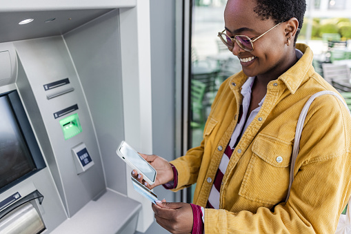 Young woman is holding a credit card and a mobile phone. She is standing in front of the ATM and thinking about the method of withdrawing money.
