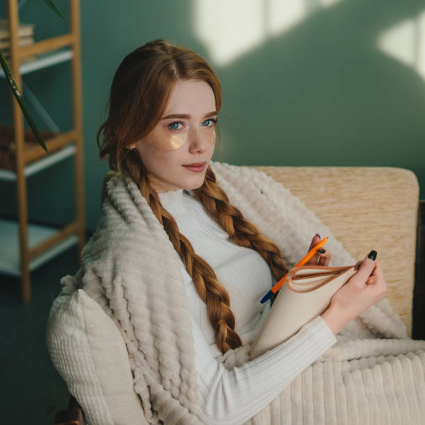 istock Ginger woman thinking and writing notes on a notebook, sitting on sofa covered with a blanket, looking at camera. House interior. 1477888015