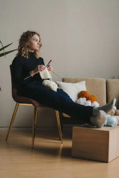 Photo of Young woman sitting in cozy armchair knitting with needles and woolen thread sitting in cozy living room. Needlework, handcraft, creative hobby or recreation.