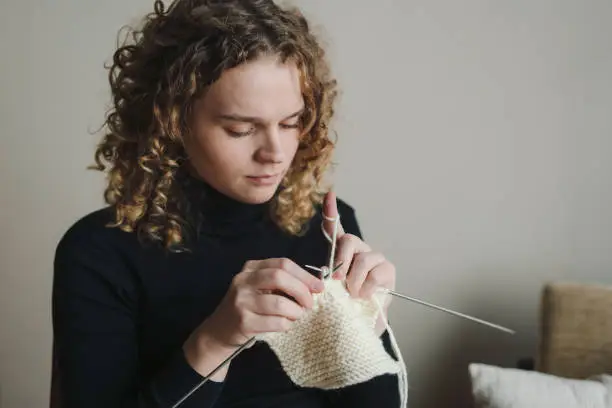 Photo of Pleasant woman sitting in living room knitting using needles, relaxing at home do favorite hobby activity on weekend. Wellbeing concept. Domestic atmosphere