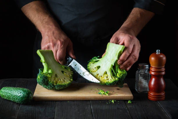 istock Professional chef cuts fresh Broccoli with a knife for salad on vintage kitchen table with fresh vegetables. Cooking and restaurant or cafe concept. 1477888338