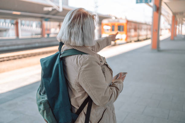 istock Old happy woman with backpack waiting train on station platform with backpack, passport and ticket shows the direction of the path on urban background. Railroad transport concept 1477888773