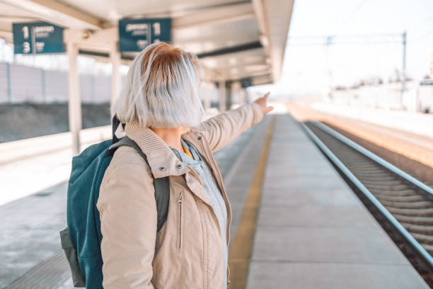 istock Old happy woman with backpack waiting train on station platform with backpack, passport and ticket shows the direction of the path on urban background. Railroad transport concept 1477889571