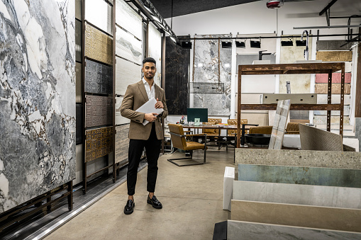 An architect in a fancy jacket and white shirt seen standing in front of some exclusive ceramic tiles and holding a laptop in a showroom during working hours.