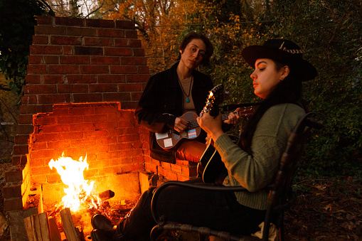 Two non-binary people play instruments by a campfire.  One of them is playing a ukulele, the other is playing a mandolin.  One is looking at their instrument, the other is looking at the other model.  They are outdoors, it is dusk in the autumn.  Both are dressed in a hip, stylish fashion.