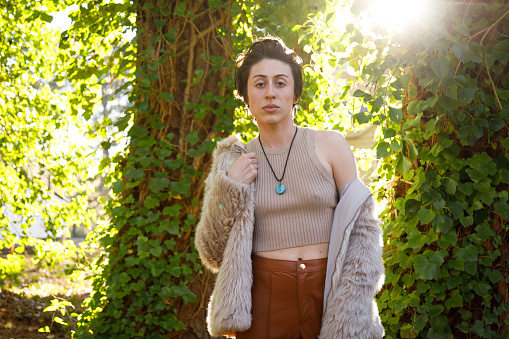 A non-binary person in front of some ivy covered trees.  They are looking at camera with a serious, confident look.    They are wearing a faux fur jacket, sleeveless top, and brown leather pants.  They have chin length hair.  They are backlit by the sun.