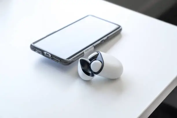 Wireless earbud headphones in their case, with the lid open, sitting on a white desk with a smartphone in the background. Room for copy space.