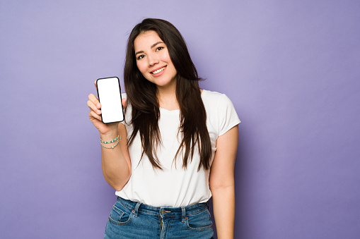 Cheerful young woman showing her smartphone screen while texting or using social media and making eye contact