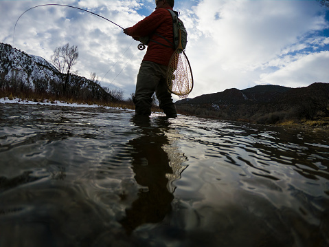Male flyfisherman in mountain stream with mountains in background from a very low angle, Flyline is very visible and in full curl