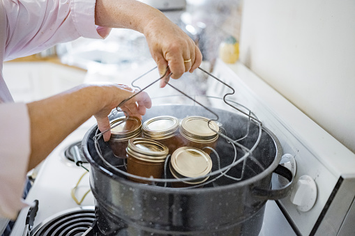 This is a close up of a senior woman’s hands sterilizing reusable glass jars in a pot steaming with boiling water on the kitchen stovetop in Miami, Florida USA.