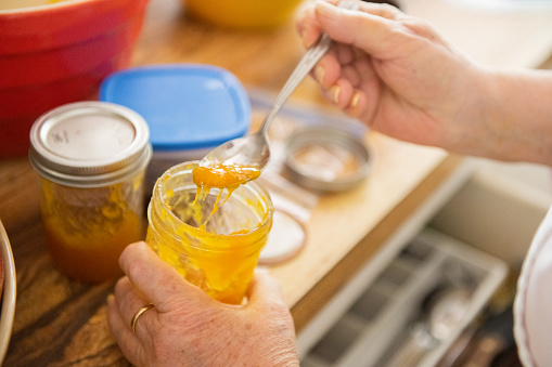 This is a close up of a senior woman’s hand spooning homemade mango jam in a glass jar in the kitchen in Miami, USA.