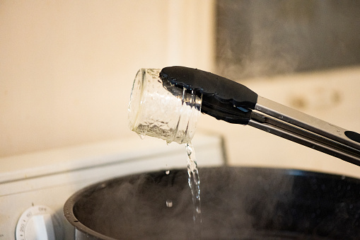 This is a close up of boiling hot water pouing out of a reusable glass jar over a pot steaming with boiling water for disinfection before canning in Miami, Florida USA.