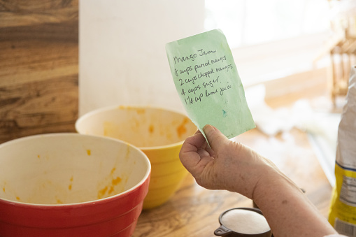 This is a photograph of a hand written recipe for mango jam being held up by the cook at home in the kitchen in Miami, Florida.