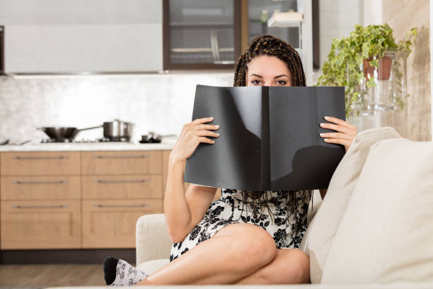Young woman on her living room sofa, with a modern kitchen in the background. Hiding her face behind a black cover booklet or notebook, her calm and captivating gaze peeks out. She has box braid hairs