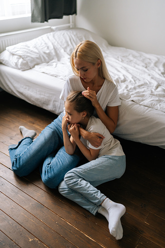 Vertical portrait of loving young mother comforting sad little child daughter, showing love and care, expressing support, hugging and stroking hair sitting on floor. Family bonding concept.