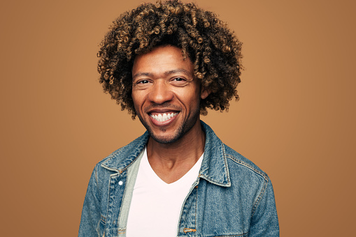 Merry young African American man in denim jacket and white t shirt with curly hair smiling and looking at camera against brown background