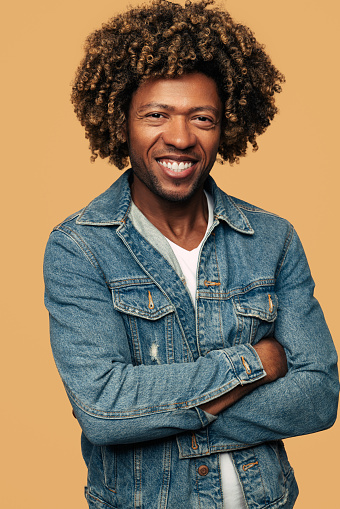 Confident young African American male model with curly dark hair in denim jacket smiling and looking at camera while standing against beige background with folded arms