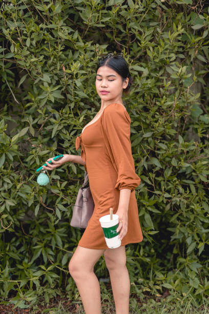 uma linda jovem olha graciosamente para baixo posando para a câmera como uma modelo com uma planta de parede atrás dela enquanto segura seu café frio e celular. - puffed sleeve - fotografias e filmes do acervo