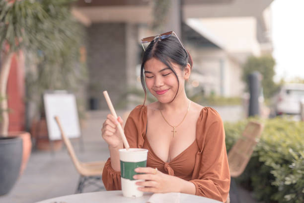 uma senhora do sudeste asiático sentada do lado de fora do café coloca a palha dentro de seu café. - puffed sleeve - fotografias e filmes do acervo