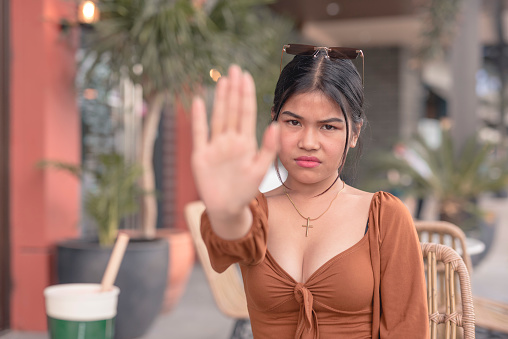 A young asian woman holds her hands in front of her face as she gestures her friend to stop talking.