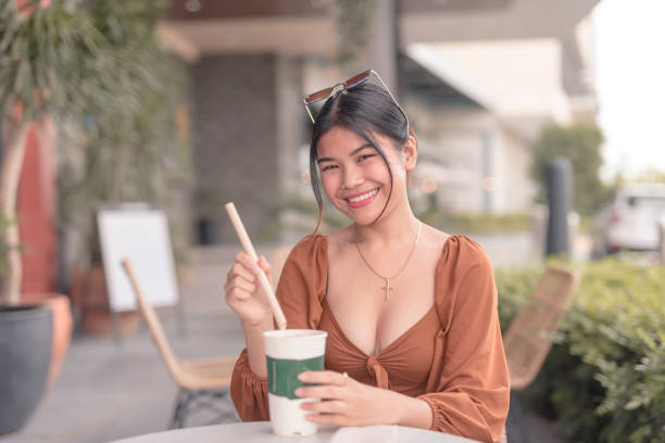 uma mulher sorridente do sudeste asiático sentada do lado de fora do café coloca a palha dentro de sua bebida. - puffed sleeve - fotografias e filmes do acervo