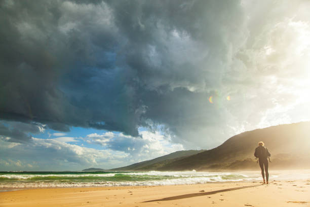 istock Young man on a journey walking along beach with bright sunlight and dark stormy clouds 1477967390