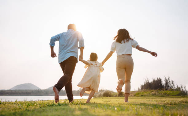 familia feliz en la luz del atardecer del parque. familia en fin de semana corriendo juntos en el prado con río los padres sostienen las manos del niño.concepto de plan de seguro de vida de salud. - family nature healthy lifestyle happiness fotografías e imágenes de stock