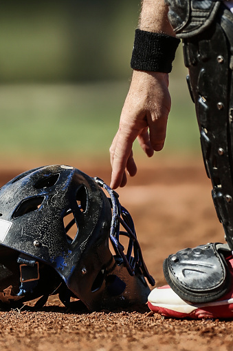 Baseball catcher reaching down to pick up facemask from the ground before the start of an inning, leg, right arm, right hand appear in the photo, closeup on mask, no faces shown in photo