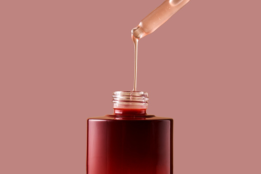 A drop of pink cosmetic drips from the dropper into a burgundy bottle.