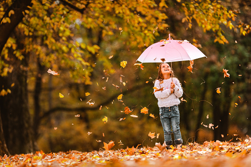 Happy little girl holding an umbrella while autumn leaves are falling onto her at the park. Copy space.