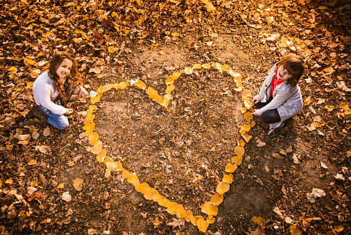 Above view of happy little couple making a heart of autumn leaves in nature and looking at camera. Copy space.