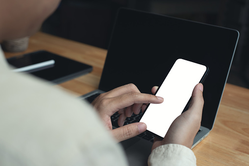 Mockup image blank white screen cell phone. Business man hand holding using mobile phone and laptop computer at home office, people communication, smartphone mobile app design