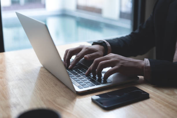 istock Businessman working on laptop computer with mobile phone on wooden table at modern office. Business man hands typing on laptop keyboard, online working 1477991672