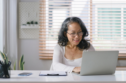 At her home office, the senior entrepreneur wears stylish glasses while working on her computer and taking notes with pen and paper
