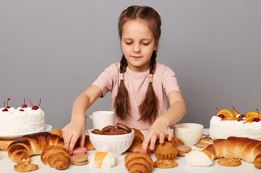 Image of serious concentrated little girl wearing casual dessert with pigtails sitting at table with delicious bakery and choosing dessert isolated over gray background