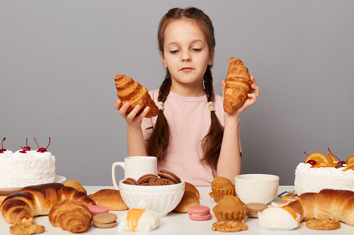 Portrait of proud happy little girl with pigtails sitting at table isolated over gray background, holding croissant in hands and looking with confident look at sweets.