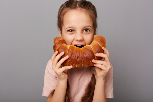 Photo of little cheerful girl with dark hair biting big yummy bagel pastry isolated over gray background, having tasty snack, eating delicious homemade bakery.
