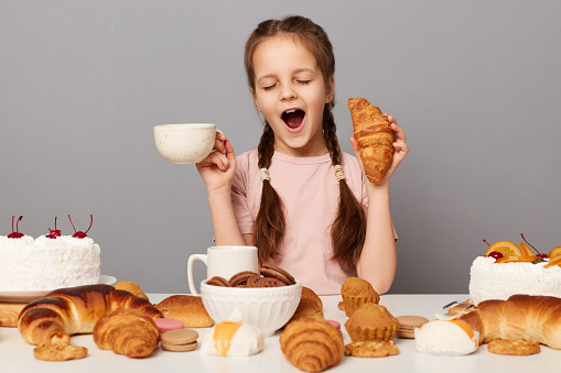 Portrait of satisfied overeating little girl with pigtails sitting at table isolated over gray background, holding cup with tea and croissant, feels sleepy, yawning.