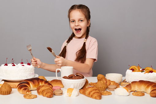 Portrait of excited hungry crazy little girl with pigtails posing near table with delicious sweets, holding knife and fork being ready to eat birthday cake isolated over gray background