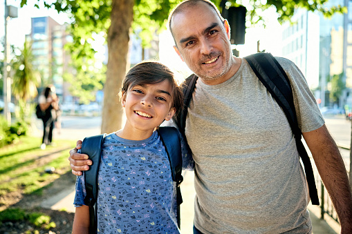Waist-up view of 11 year old boy with his 45 year old father, both wearing casual clothing and backpacks, grinning at camera with sunshine in background.