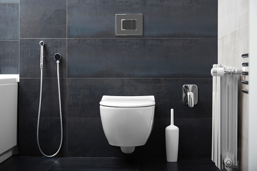 Modern wall-mounted white toilet bowl, chrome flush button and bidet hygienic shower against the background of a black bathroom wall. Part of the interior of the bathroom in the apartment.